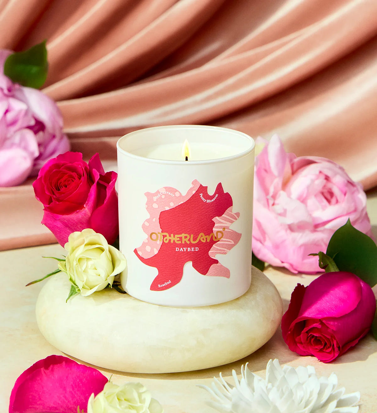 Candle with a decorative label surrounded by pink and white roses on a soft pink fabric background