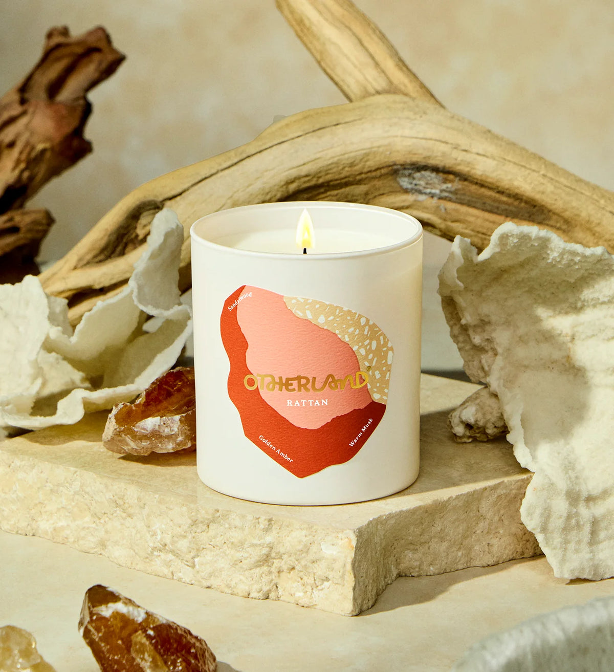 Candle with 'Otherland' branding on a natural setting with driftwood and shells.