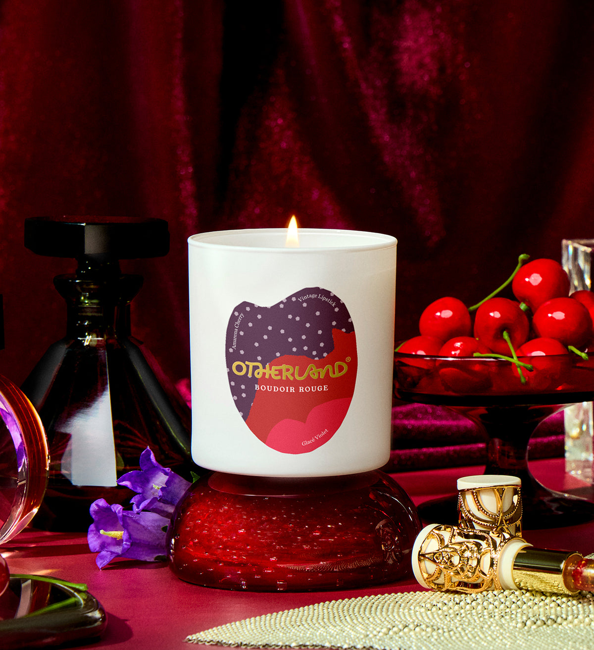 Lit candle with 'Otherland' branding on a red surface with cherries and perfume bottles.