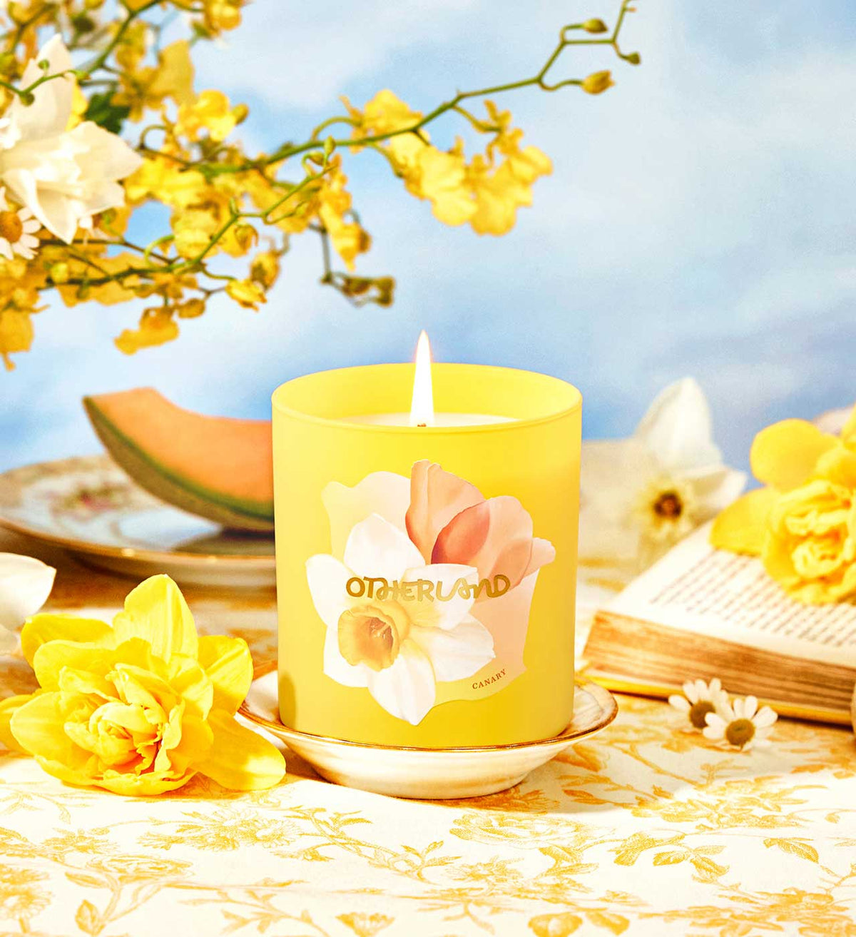 Yellow candle with floral design on a table with yellow flowers and a blue sky background