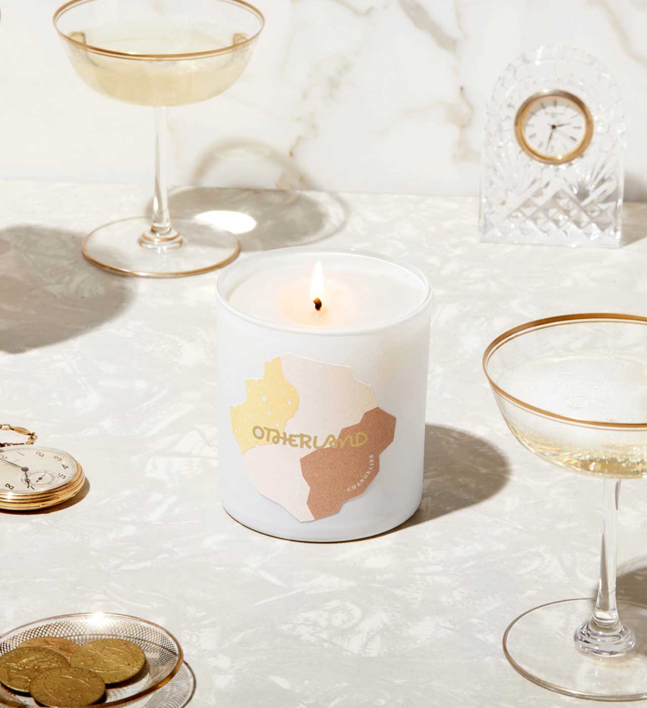 Candle with 'Otherland' branding on a table with glasses and coins