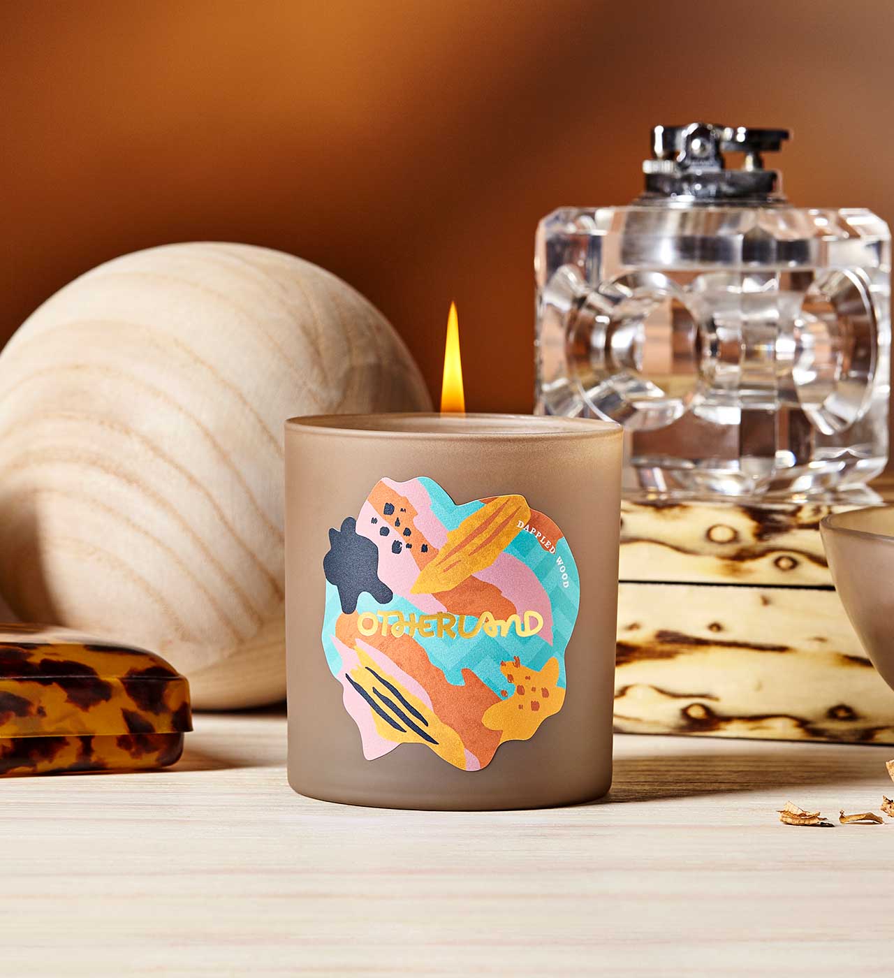 Candle with colorful design and 'Otherland' branding on a wooden surface.