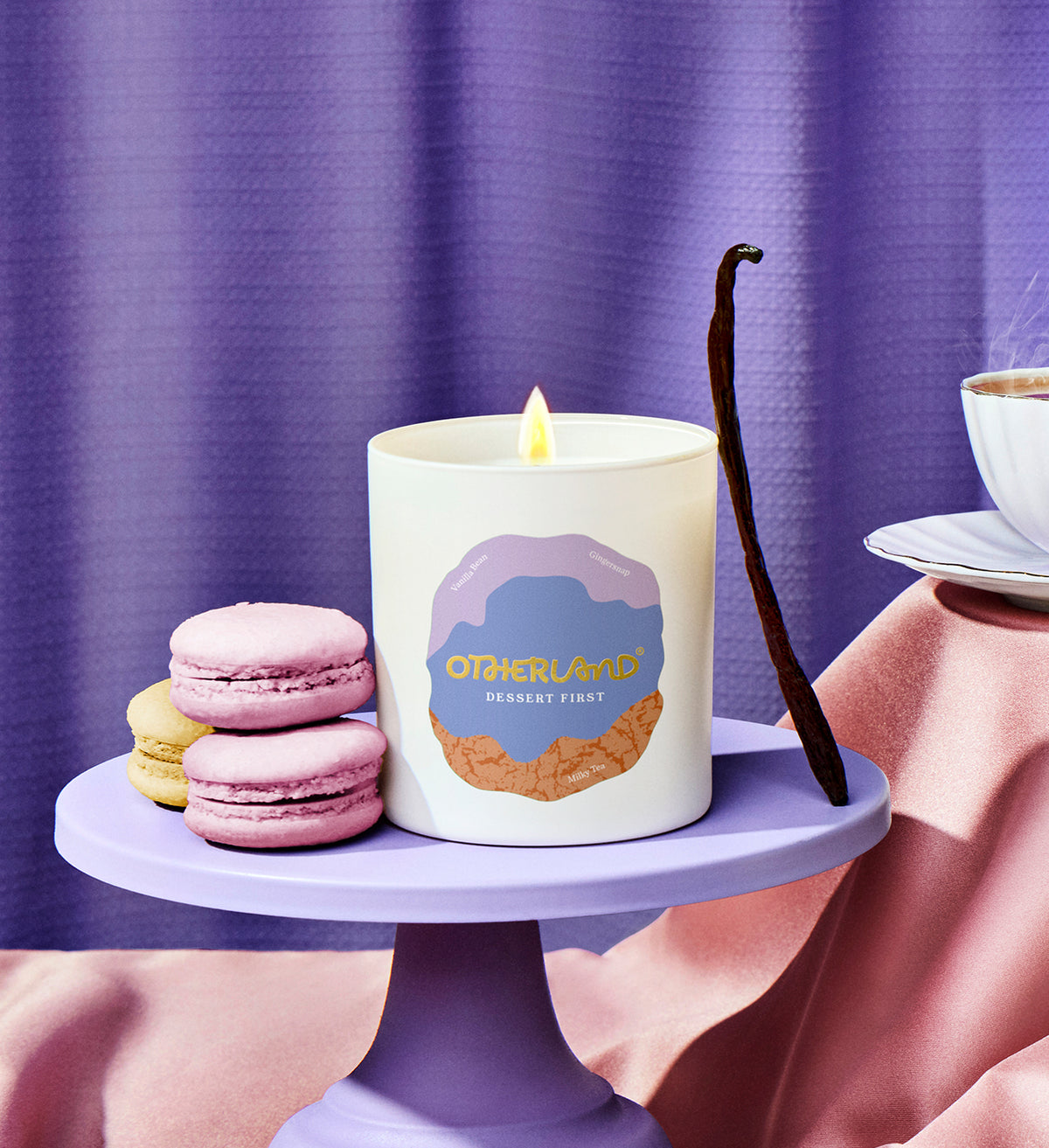 Candle with a dessert design on a purple stand with macarons and a vanilla bean against a purple curtain.