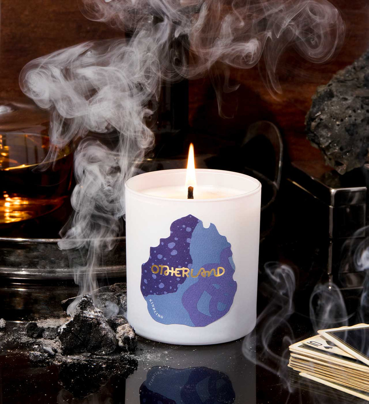 Candle with 'Otherland' logo on a dark background with smoke and rocks.