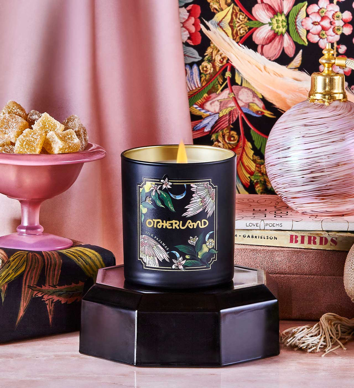 Black candle with gold lid on a decorative stand, surrounded by a pink lamp, books, and a colorful floral pattern.