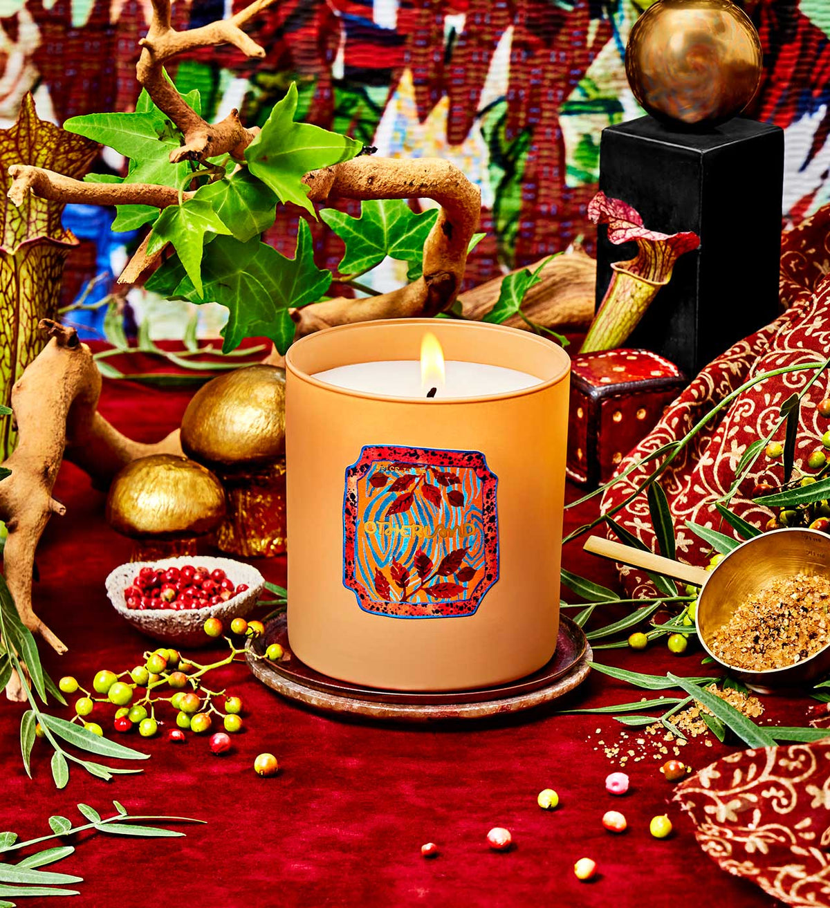Decorative candle with intricate design on a red surface with plants and small objects.